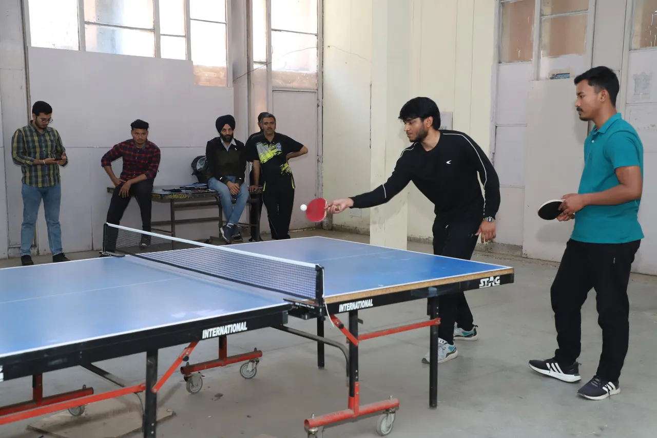 A table tennis player stretches over the table while a partner watches