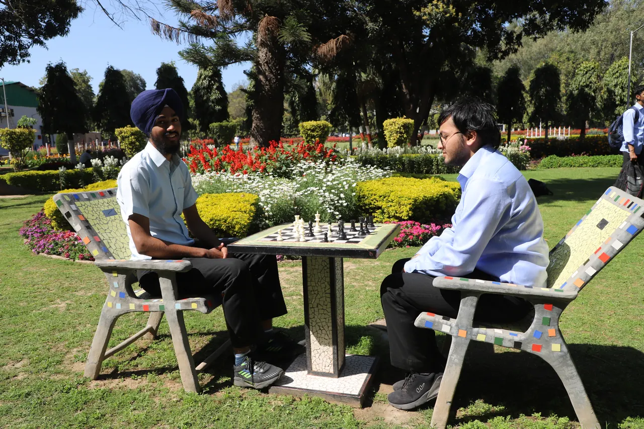 Two players face each other over an outdoor chess table in a garden