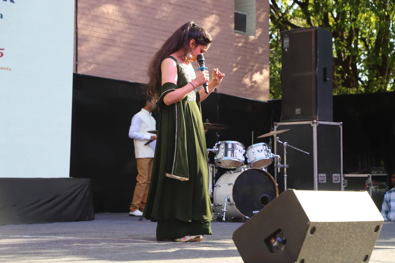 A performer holds a microphone on the outdoor stage with a drum kit behind her