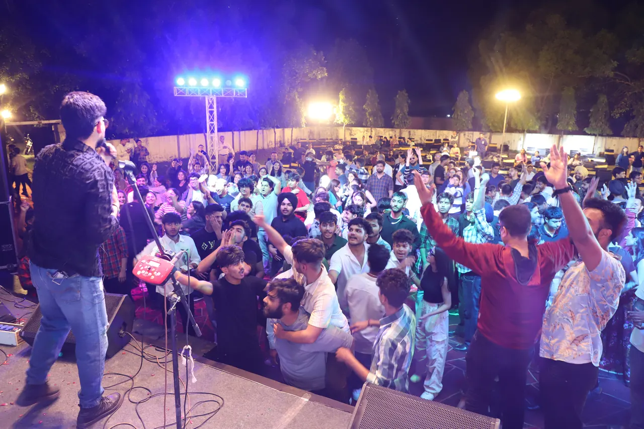 A singer performs on an outdoor stage at night as a large crowd raises their arms under floodlights