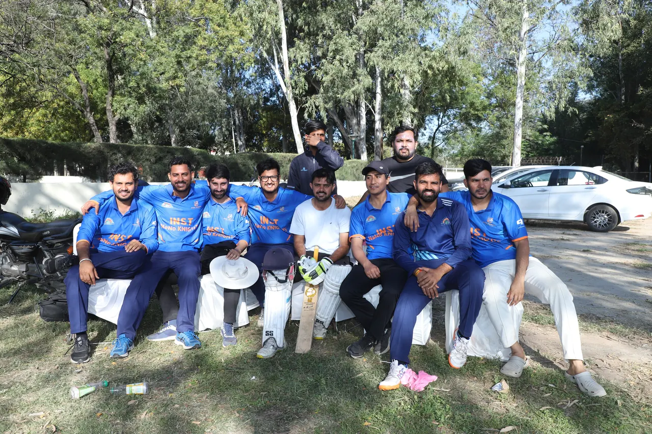 Cricket players in blue jerseys sit together beside the ground