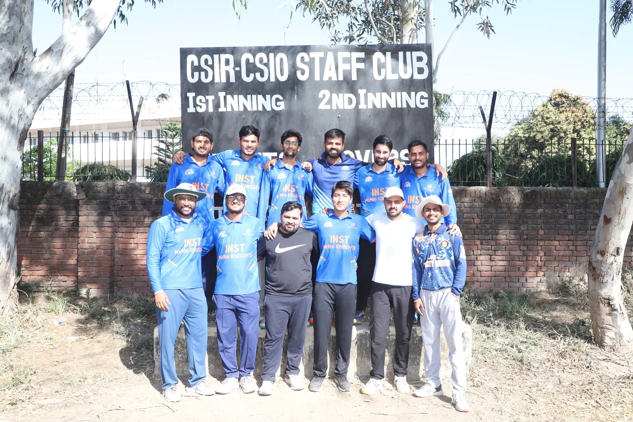 A cricket team in blue poses in front of the Staff Club scoreboard
