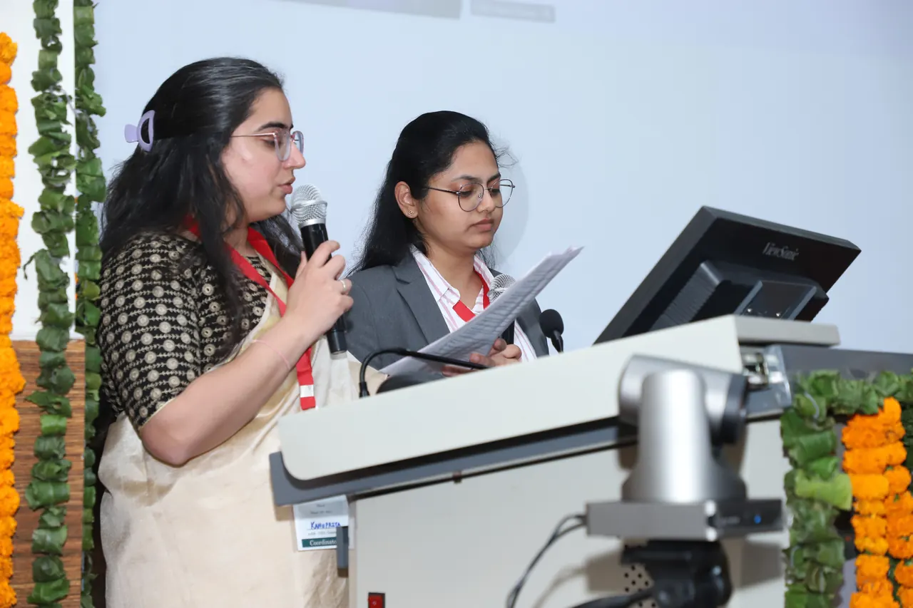 Two hosts speak from the podium in an auditorium decorated with marigolds